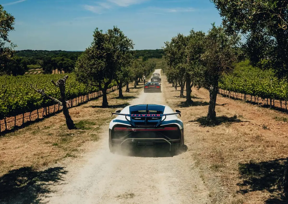 Kolona superšportnikov Bugatti – vodilni bel Bugatti dviguje prah na makadamski poti med vinogradi in oljkami v portugalskem podeželju. / Convoy of Bugatti hypercars, lead white Bugatti kicking up dust on a gravel lane flanked by vineyards and olive trees in the Portuguese countryside.