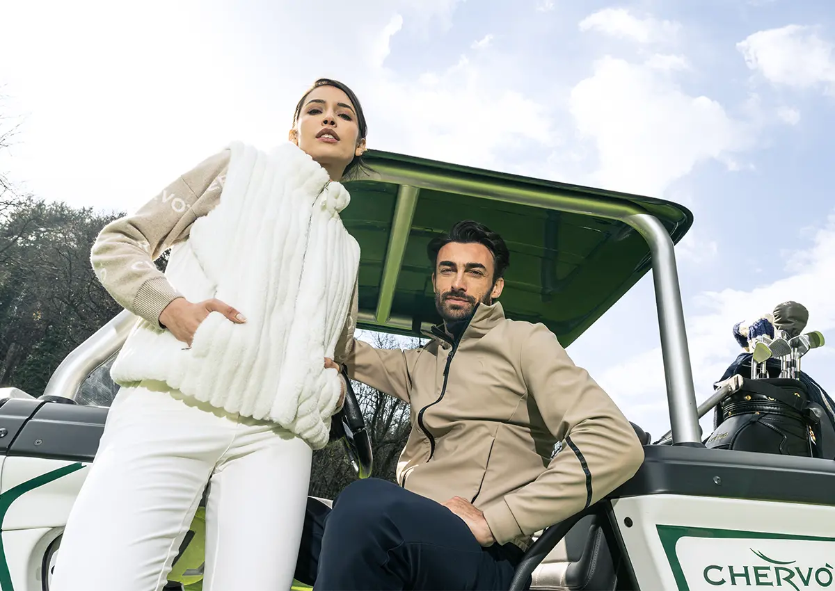 Moški in ženska v športno elegantnih oblačilih Chervò iz kolekcije jesen–zima 2025 stojita ob golf vozičku na igrišču.