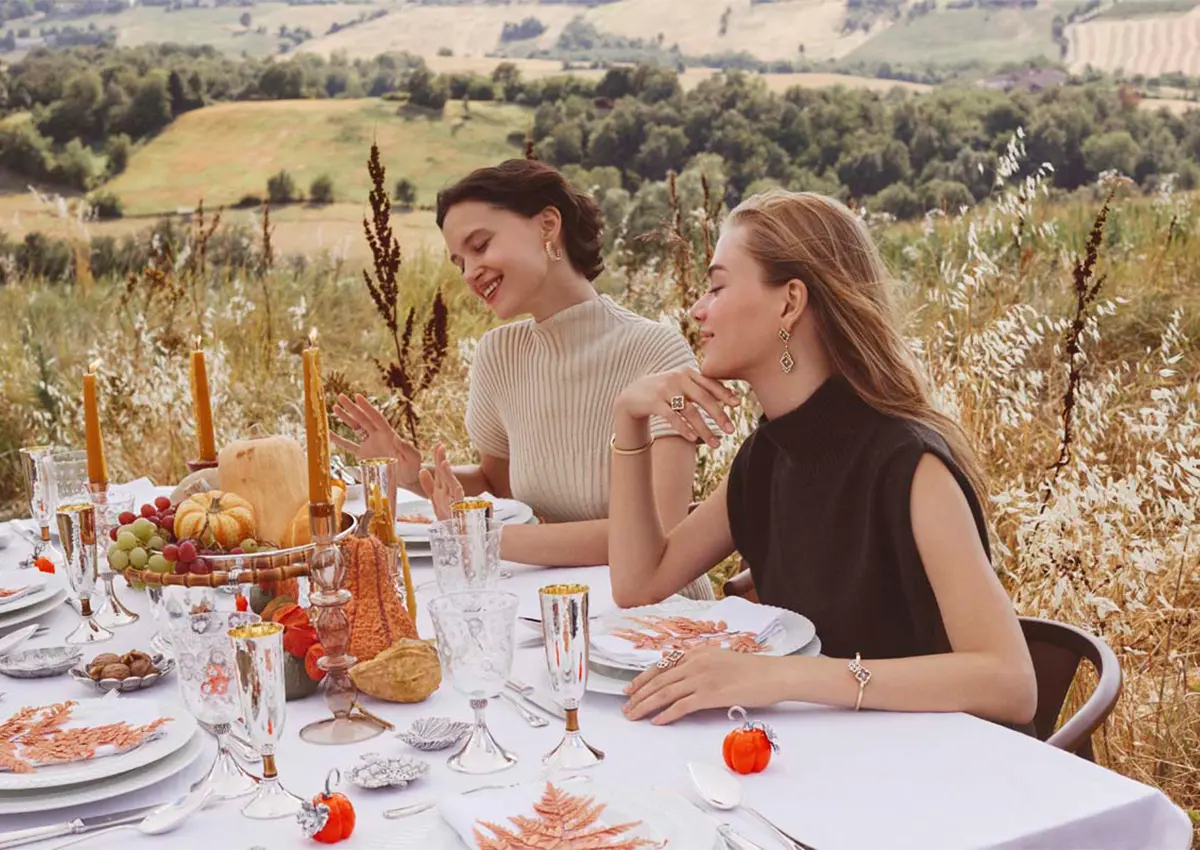 Dve elegantno oblečeni ženski uživata ob jesensko okrašeni mizi z izdelki Buccellati iz srebra in Murano stekla, obdani s toplimi barvami narave.