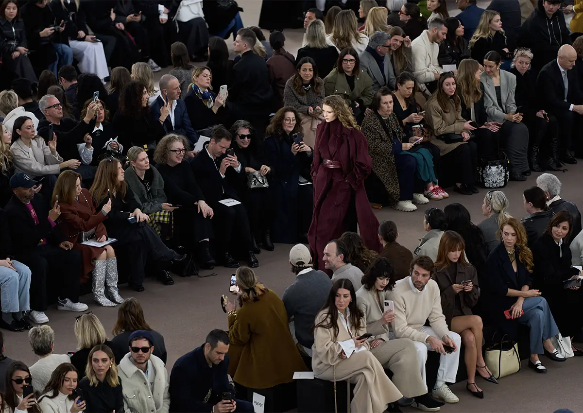 Modna revija Max Mara Fall/Winter 2025, model v dolgstem rdečem plašču hodi po pisti med občinstvom, ki spremlja novo kolekcijo.