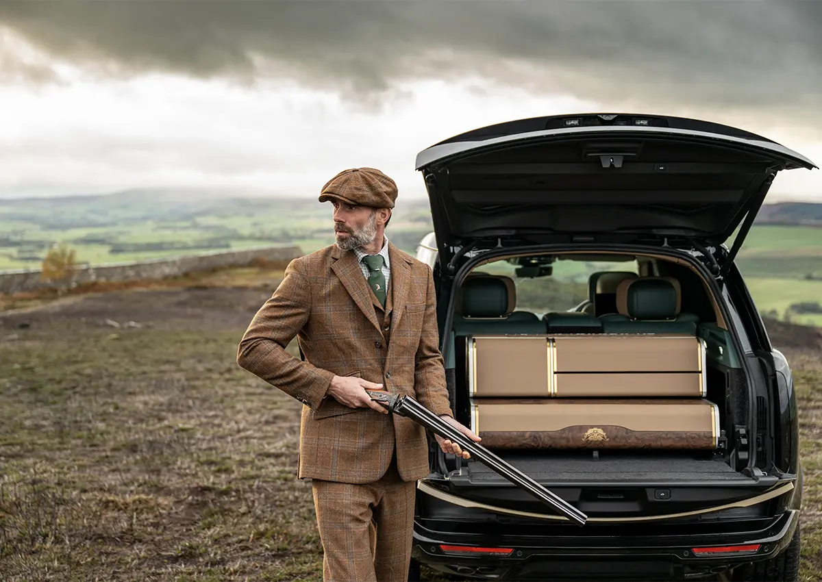 Moški v klasični karirani obleki stoji ob odprtem prtljažniku Range Roverja Holland & Holland Edition by Overfinch, v rokah drži lovsko puško, v ozadju pa se razprostira podeželska pokrajina.